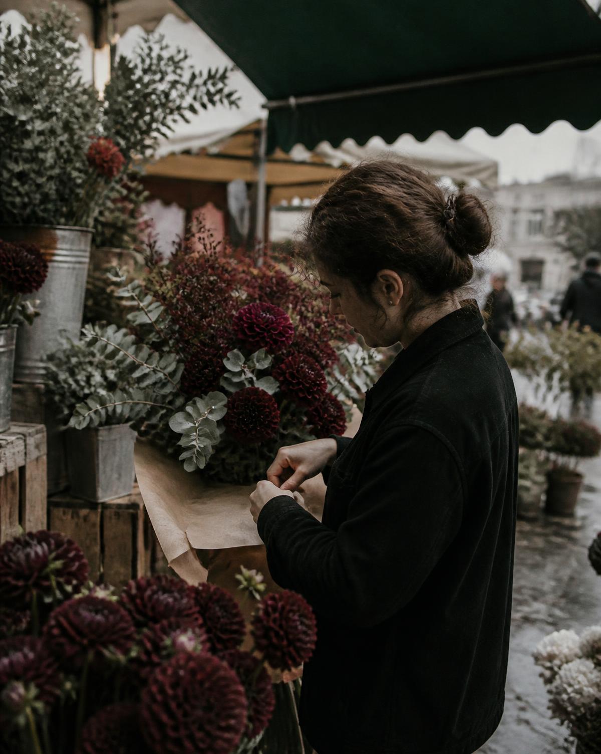 A florist wrapping burgundy dahlias and eucalyptus in brown paper at a market stall